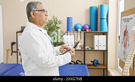 Un uomo maturo, probabilmente un medico, studia una compressa all'interno di una clinica di riabilitazione illuminata, circondata da poster e attrezzature mediche. Foto Stock