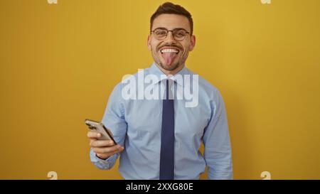 Un bel giovane ispanico con la barba e gli occhiali si attacca sulla lingua mentre tiene in mano un telefono, isolato su un vibrante sfondo giallo. Foto Stock