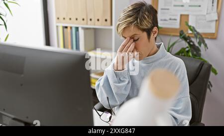 Donna ispanica stressata con capelli biondi corti che sente mal di testa in un moderno ambiente d'ufficio. Foto Stock