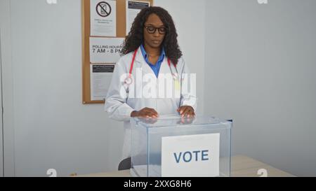 Una donna afroamericana adulta con i capelli ricci, vestita con il cappotto di un medico, getta il suo voto al chiuso in una stazione elettorale. Foto Stock