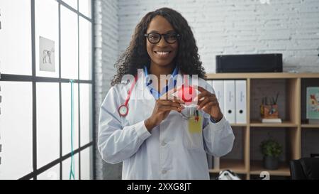 Una donna afroamericana in un cappotto bianco, che tiene un cuore rosso nella sua clinica veterinaria, sorride con fiducia con capelli ricci e occhiali. Foto Stock