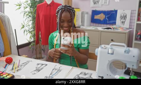 Una donna africana sorridente con trecce che utilizza uno smartphone in un colorato atelier di sartoria pieno di schizzi alla moda e attrezzature da cucito. Foto Stock