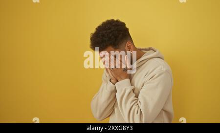 Un uomo in difficoltà copre il suo volto con le mani su uno sfondo giallo, trasmettendo emozioni e disperazione. Foto Stock