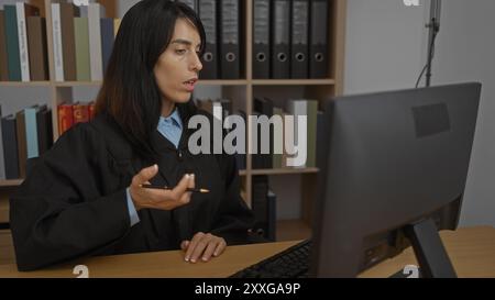 Giudice donna che lavora in ufficio, indossa l'abito, davanti al computer, tiene una matita, libreria in background, discute, giovane adulto che si concentra sul legale Foto Stock