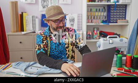 Uomo con barba in camicia colorata che usa un computer portatile in un creativo negozio di sartoria con macchina da cucire e bobine di filo Foto Stock