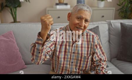 Senior ispanico con capelli grigi in camicia a quadri, sorridente mentre si siede su un divano in un accogliente soggiorno. Foto Stock