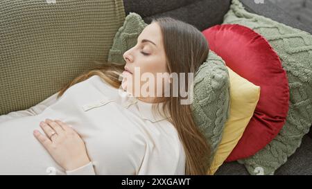 Una giovane donna incinta ispanica che dorme tranquillamente a casa su un divano con cuscini colorati. Foto Stock