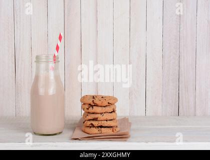 Uno spuntino a base di biscotti e latte su un set di legno rustico. Una piccola bottiglia di latte al cioccolato con cannuccia da bere e una pila di biscotti con scaglie di cioccolato. Oriz Foto Stock