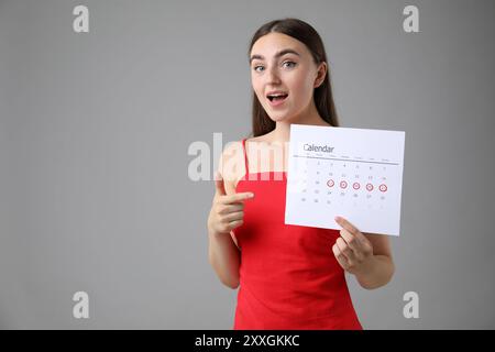 Donna che tiene il calendario con giorni di ciclo mestruale contrassegnati su sfondo grigio, spazio per il testo Foto Stock