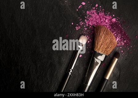 Make-up pennelli con cosmetici frantumato, girato dalla parte superiore su uno sfondo nero con copia spazio, una bellezza modello di progettazione per un banner per il trucco Foto Stock