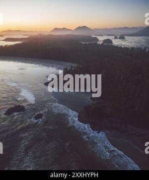 Una costa panoramica al tramonto con onde che si infrangono sulla spiaggia, circondata da montagne e foreste sotto un cielo nuvoloso Tofino, BC, Canada, Nord America Foto Stock