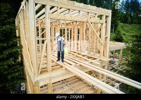 Lavoratore costruendo casa telaio di legno vicino alla foresta. Uomo che tratta legno, applicando ignifugo con spruzzatore, mentre vestito con tuta protettiva, casco. Concetto di moderna costruzione ecologica. Foto Stock