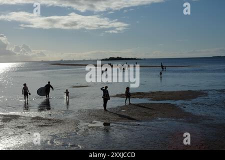 Persone che camminano indietro attraverso l'acqua di mare da King Island con la bassa marea a Wellington Point, in sagoma contro una penisola sull'acqua di Morton Bay Foto Stock