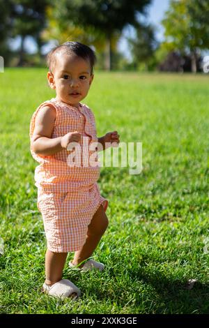 Una bambina latina di un anno che cammina da sola in un parco guardando la macchina fotografica Foto Stock