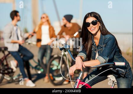 Pronti a divertirsi e ad avventurarsi. Bella giovane donna sorridente che si appoggia alla sua bicicletta e guarda la fotocamera mentre i suoi amici parlano in sottofondo Foto Stock