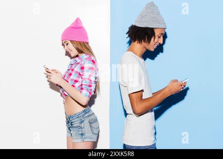 Gli opposti si attraggono. Giovane coppia funky che tiene i telefoni cellulari e si mette dietro dietro mentre si sta in piedi su uno sfondo colorato Foto Stock