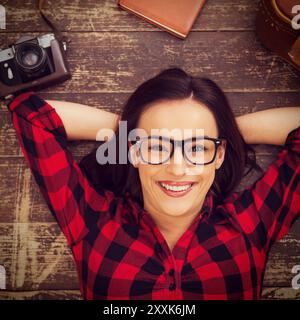 Sognando un grande viaggio. Vista dall'alto di una bellissima giovane donna con occhiali sdraiata sul pavimento in legno e sorridente mentre la valigia e la macchina fotografica sono stese vicino a lei Foto Stock