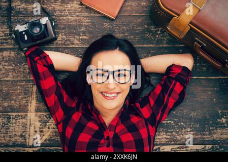 Pronti per un grande viaggio. Vista dall'alto di una bellissima giovane donna con occhiali sdraiata sul pavimento in legno e sorridente mentre la valigia e la macchina fotografica sono stese vicino a lei Foto Stock