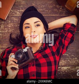 Fotografo creativo. Vista dall'alto di una bella giovane donna con i copricapo sdraiata sul pavimento e con la fotocamera mentre la valigia e il blocco note si appoggiano vicino a lei Foto Stock