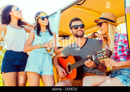 Passare del tempo di qualità con gli amici. Bel giovane che siede al minivan e suona la chitarra mentre tre ragazze gli stanno vicino e sorridono Foto Stock