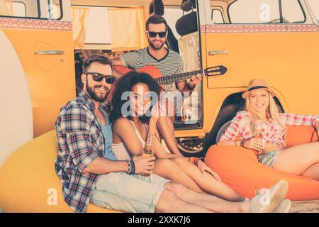 Passare l'estate insieme. Tre giovani felici seduti vicino al loro minivan con bottiglie di birra mentre l'uomo suona la chitarra Foto Stock