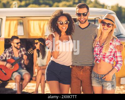 Passare del tempo con gli amici. Giovane felice che abbraccia due belle donne mentre giovane coppia seduto in un minivan retrò sullo sfondo Foto Stock