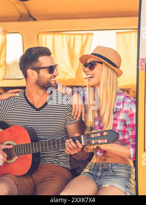 Passare del tempo libero insieme. Bel giovane uomo seduto in minivan e che suona la chitarra mentre la sua ragazza si lega a lui e sorride Foto Stock