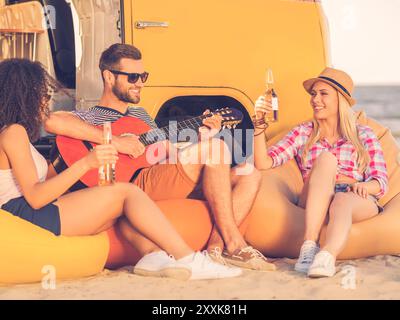 Passare del tempo con gli amici. Giovane felice che suona la chitarra mentre due giovani donne sedute vicino a lui e bevono birra con un minivan giallo in t Foto Stock