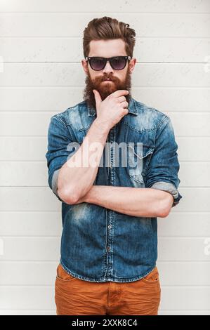 Fresco e sicuro di sé. Giovane uomo barbuto che guarda la macchina fotografica e tiene la mano sul mento mentre si trova all'aperto Foto Stock