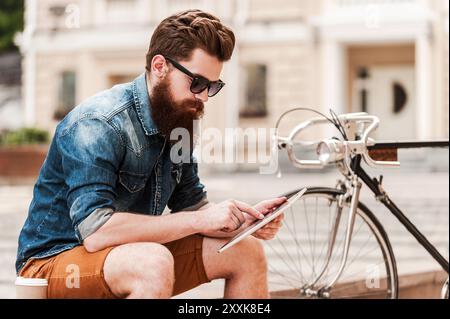 Sto recuperando qualche notizia. Bel giovane barbuto che tiene il tavolo digitale seduto vicino alla sua bicicletta all'aperto Foto Stock