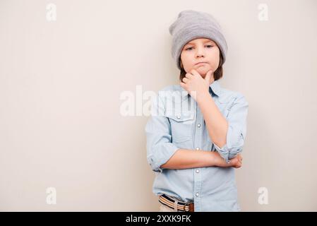 Nel profondo dei suoi pensieri. Bel ragazzino in cappello che tiene per mano il mento e guarda la macchina fotografica mentre è in piedi su sfondo grigio Foto Stock