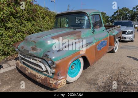Rayleigh, Regno Unito. 25 agosto 2024. Un pick-up vintage molto resistente alle intemperie con carrozzeria arrugginita e vernice sbiadita si trova accanto a un moderno SUV argentato. Le ruote anteriori turchesi e i parafanghi arrotondati del veicolo evocano il design della metà del XX secolo, in netto contrasto con il veicolo contemporaneo e lo sfondo urbano soleggiato. Auto e camion americani si incontrano al garage Farm Fresh. L'evento consente ai proprietari di portare con sé i propri veicoli in modo che i visitatori possano osservare da vicino. Penelope Barritt/Alamy Live News Foto Stock