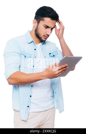 Di che si tratta? Frustrato giovane indiano che tiene la mano nei capelli mentre guarda il suo tablet digitale e si trova su sfondo bianco Foto Stock