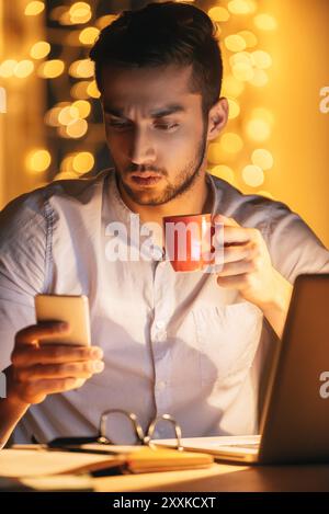 Non ho tempo per una pausa. Giovane sicuro di sé che guarda il suo smartphone e tiene in mano una tazza di caffè mentre è seduto sul posto di lavoro di notte con Foto Stock