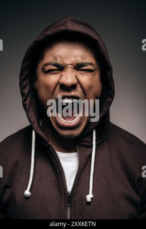 Energia dentro di lui. Furioso giovane africano in camicia con cappuccio che tiene gli occhi chiusi e grida mentre si trova su sfondo grigio Foto Stock