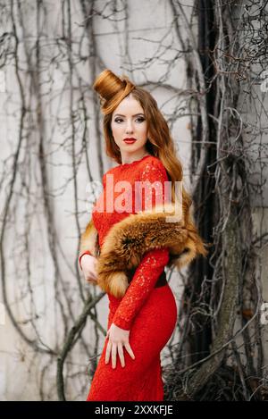 ragazza giovane con capelli rossi in un abito rosso brillante vicino ad un albero in un parco vuoto Foto Stock