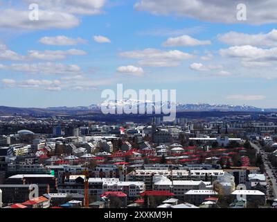 Una vista sopraelevata di una vivace città adornata da tetti rossi, adagiata sullo sfondo di lontane montagne innevate e di un cielo blu parzialmente nuvoloso Foto Stock