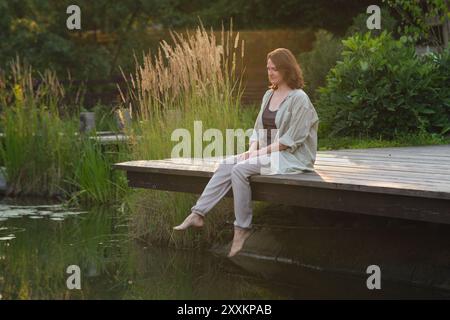 Donna seduta sul bordo di un ponte di legno vicino a uno stagno circondato da erba alta e vegetazione. Fotografia di ritratti per esterni per design e stampa. Relaxat Foto Stock