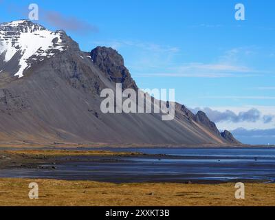 Questa immagine mostra un'aspra montagna con cime innevate, un primo piano di sabbia piatta e prato, che si estende fino all'oceano calmo sotto un cielo blu brillante. Foto Stock