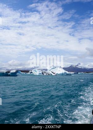 L'immagine mostra splendidi iceberg che galleggiano in una tranquilla laguna glaciale sotto un cielo blu brillante, con montagne maestose sullo sfondo coperte Foto Stock