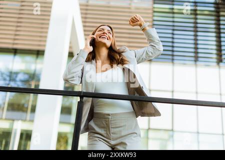 Una donna d'affari gioiosa festeggia un momento all'aperto, partecipando a una allegra conversazione telefonica mentre si appoggia a una ringhiera accanto a un elegante buildi Foto Stock