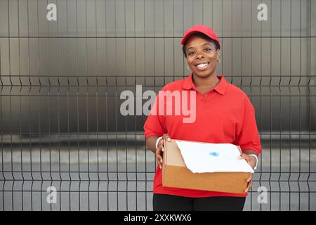 Il lavoratore che sorride tenendo la confezione marrone davanti alla recinzione in metallo, vestito con uniforme rossa con berretto coordinato e pantaloni cachi, offrendo un servizio professionale Foto Stock