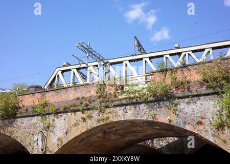 Manchester, Greater Manchester, Regno Unito. 24 agosto 2024: Ponte ferroviario industriale su un vecchio arco in mattoni ricoperto di verde sotto cieli azzurri. Foto Stock