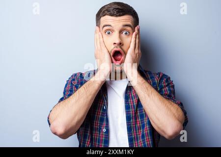 Mi sento scioccato. Giovane frustrato con una camicia casual che tiene la bocca aperta e sembra terrorizzato toccando il viso e in piedi contro il dorso grigio Foto Stock