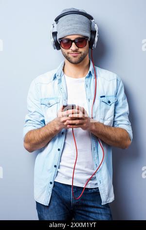 Sto cercando la sua canzone preferita. Bel giovane ed elegante uomo con le cuffie che reggono il lettore MP3 e lo guardano mentre è in piedi su uno sfondo grigio Foto Stock