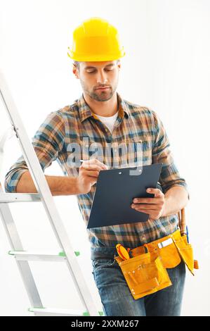 Maestro di costruzione al lavoro. Giovane costruttore serio che scrive negli appunti e si appoggia alla scala mentre è in piedi all'interno Foto Stock