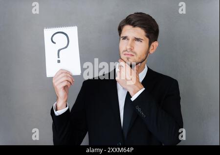 In cerca di risposta. Giovane uomo d'affari con un blocco note con un punto interrogativo su di esso e con lo sguardo rivolto verso l'esterno mentre si trova su sfondo grigio Foto Stock