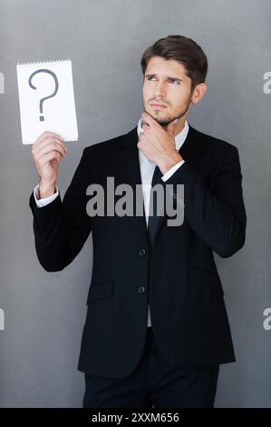 Ha una grande domanda. Giovane uomo d'affari con un blocco note con un punto interrogativo su di esso e con lo sguardo rivolto verso l'esterno mentre si trova su sfondo grigio Foto Stock