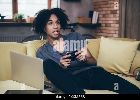 Giocatore uscito. Giovane africano eccitato che gioca ai videogiochi e tiene la bocca aperta mentre si siede sul divano di casa Foto Stock