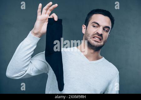 Cos'è quello?! Giovane frustrato che esprime negatività e copre il naso con le dita tenendo il calzino nero e in piedi su sfondo grigio Foto Stock